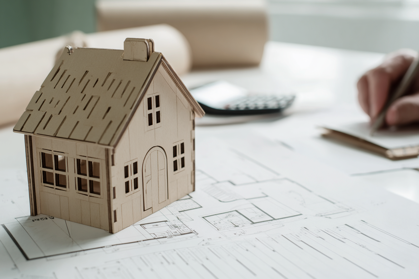 Maquette de maison en bois posée sur un plan architectural avec une main écrivant en arrière-plan