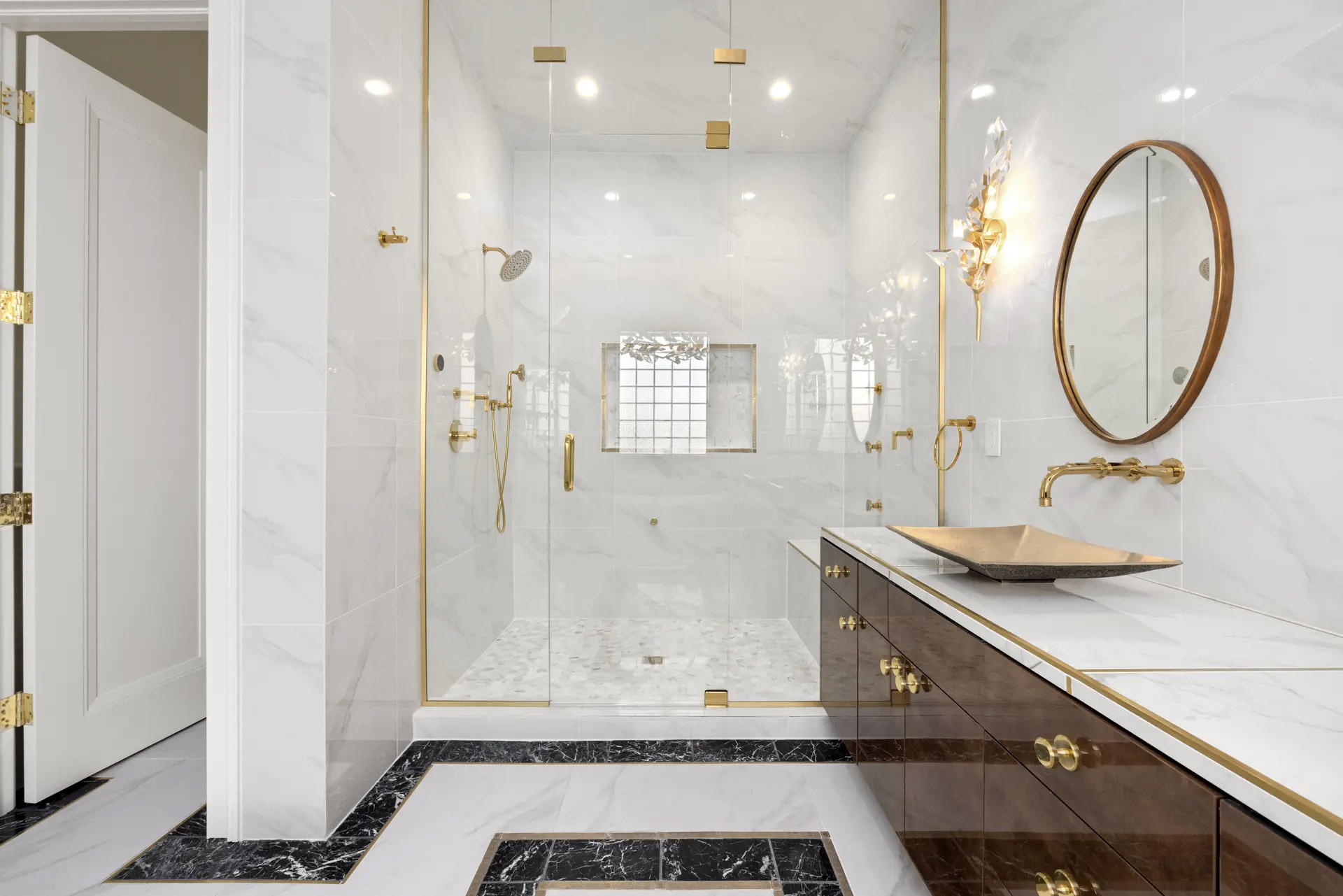 Salle de bain luxueuse et lumineuse avec douche vitrée en marbre blanc, robinetterie dorée, meuble-lavabo en bois foncé, vasque décorative, miroir rond et éclairage mural élégant