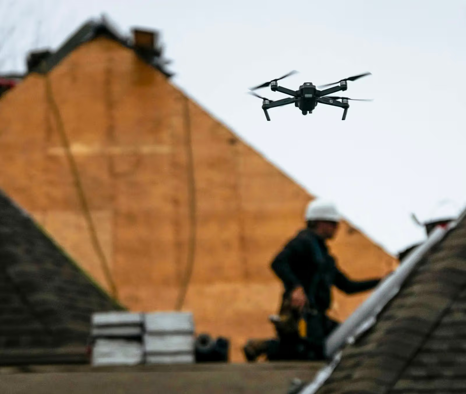 Drone en vol au-dessus d’un chantier de toiture, avec couvreur flou en arrière-plan, bardeaux, matériaux et charpente en bois.