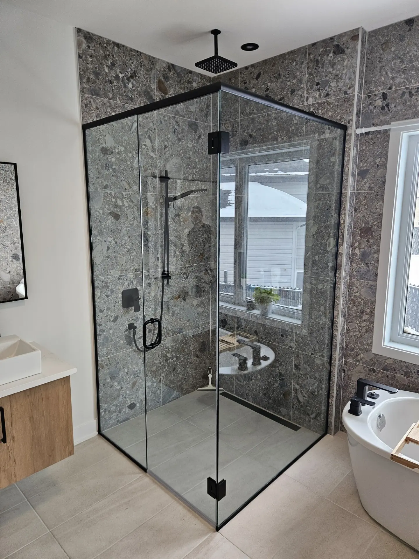 Modern glass shower with a black frame, terrazzo-effect tiles, black fixtures, and a rainfall shower in a bright bathroom with a freestanding tub.