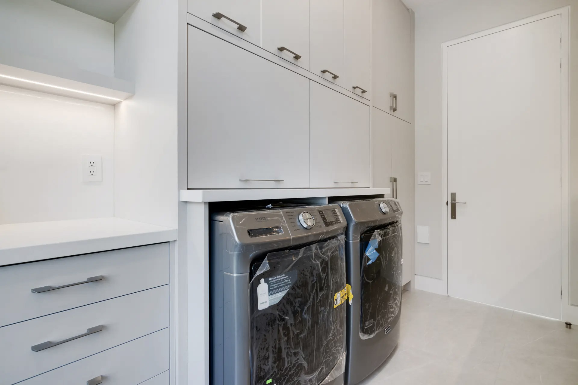 Modern bright laundry room with gray washer and dryer, full-height white cabinets, integrated countertop, storage drawers, metal handles, under-cabinet lighting and beige ceramic floor
