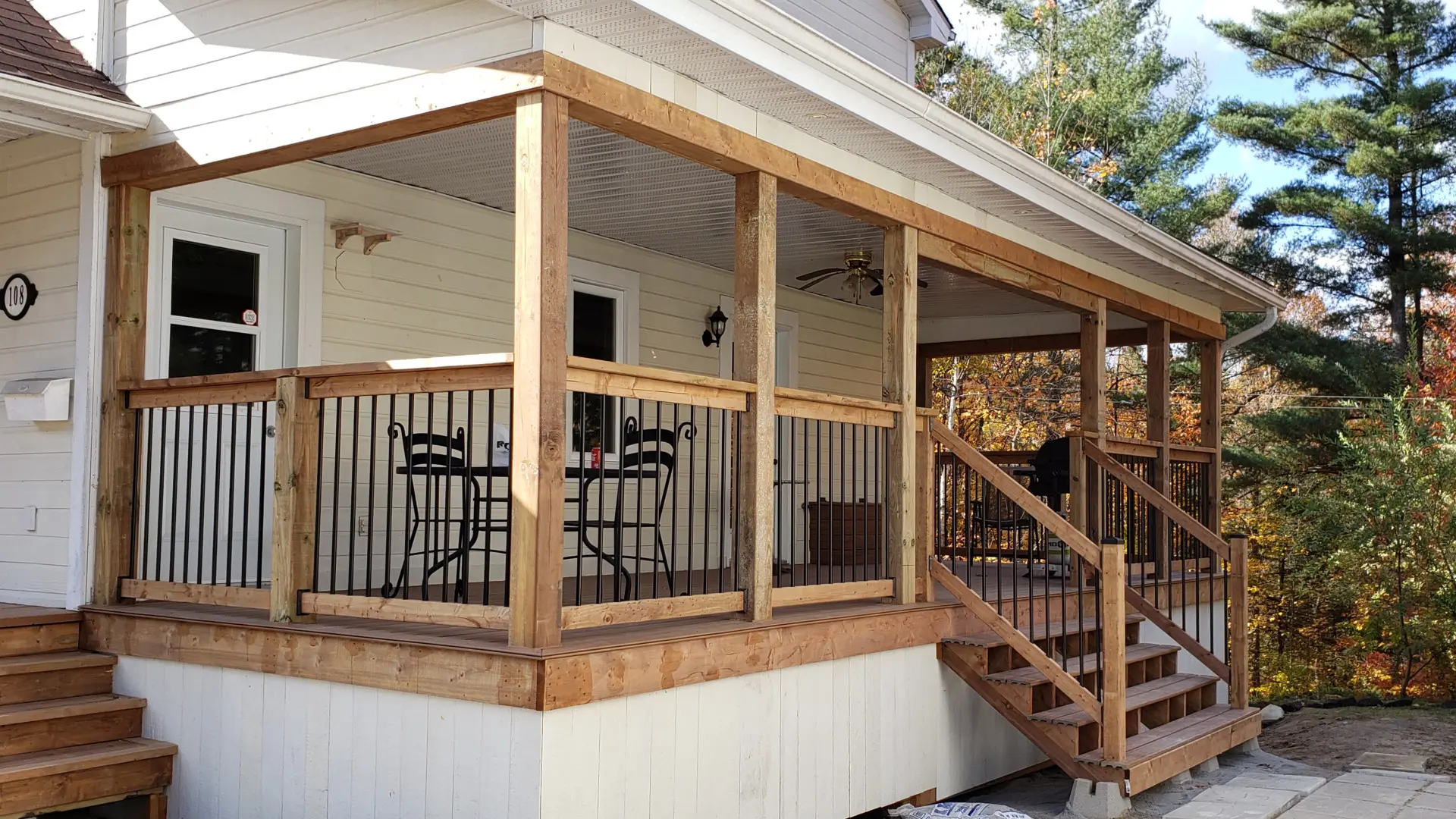 Covered wood porch with posts, black metal railings, stairs, and a deck attached to a house with light-colored siding