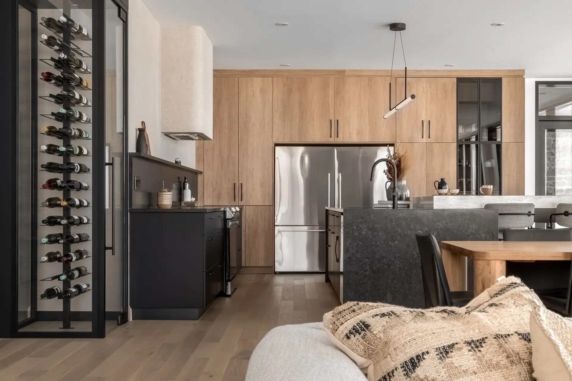 Cuisine moderne ouverte avec armoires en bois, îlot en pierre, cellier vitré et supports à bouteilles noirs