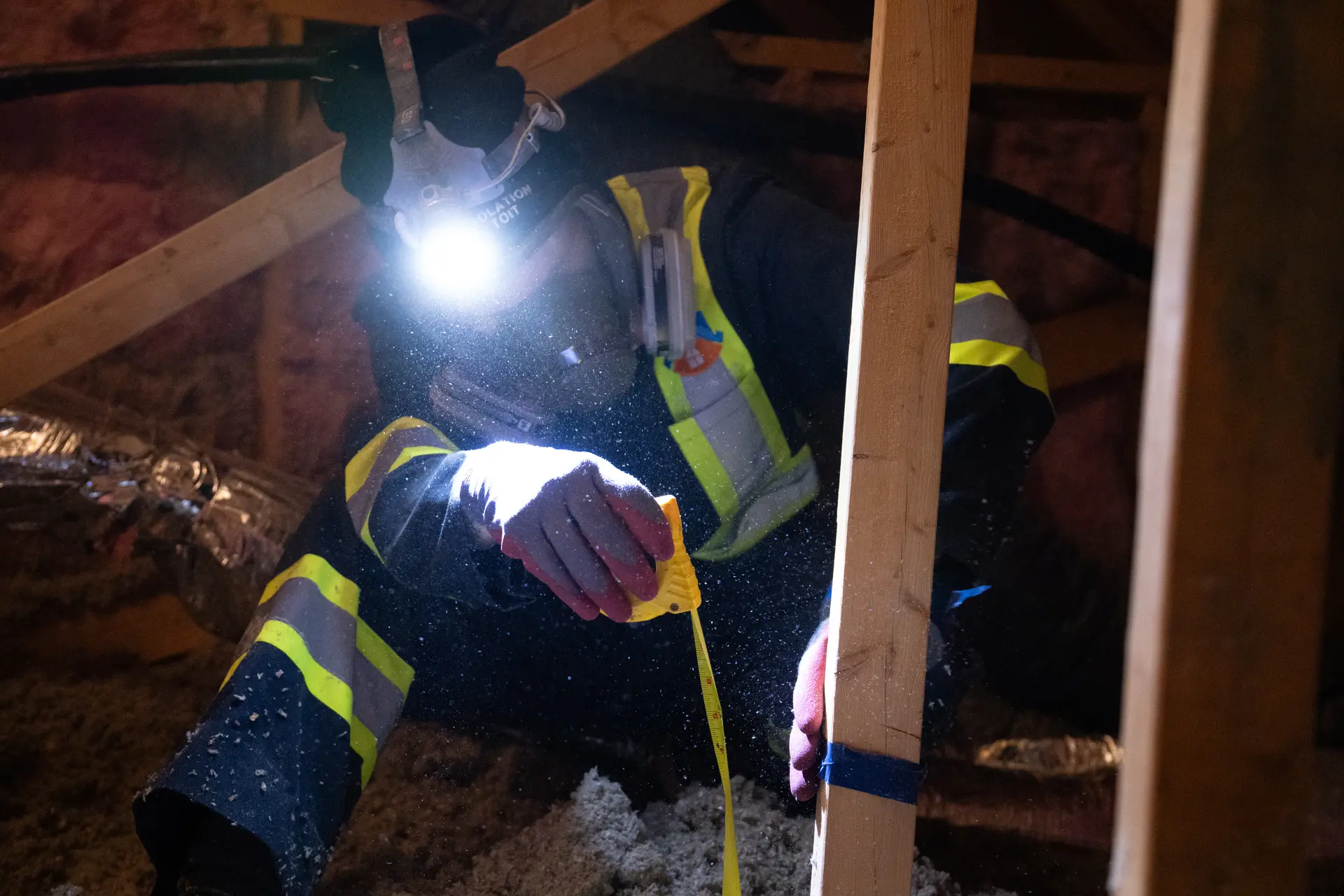 Travailleur effectuant une inspection dans un entretoit avec lampe frontale, ruban à mesurer et équipement de protection pour évaluer l’isolation et la structure.