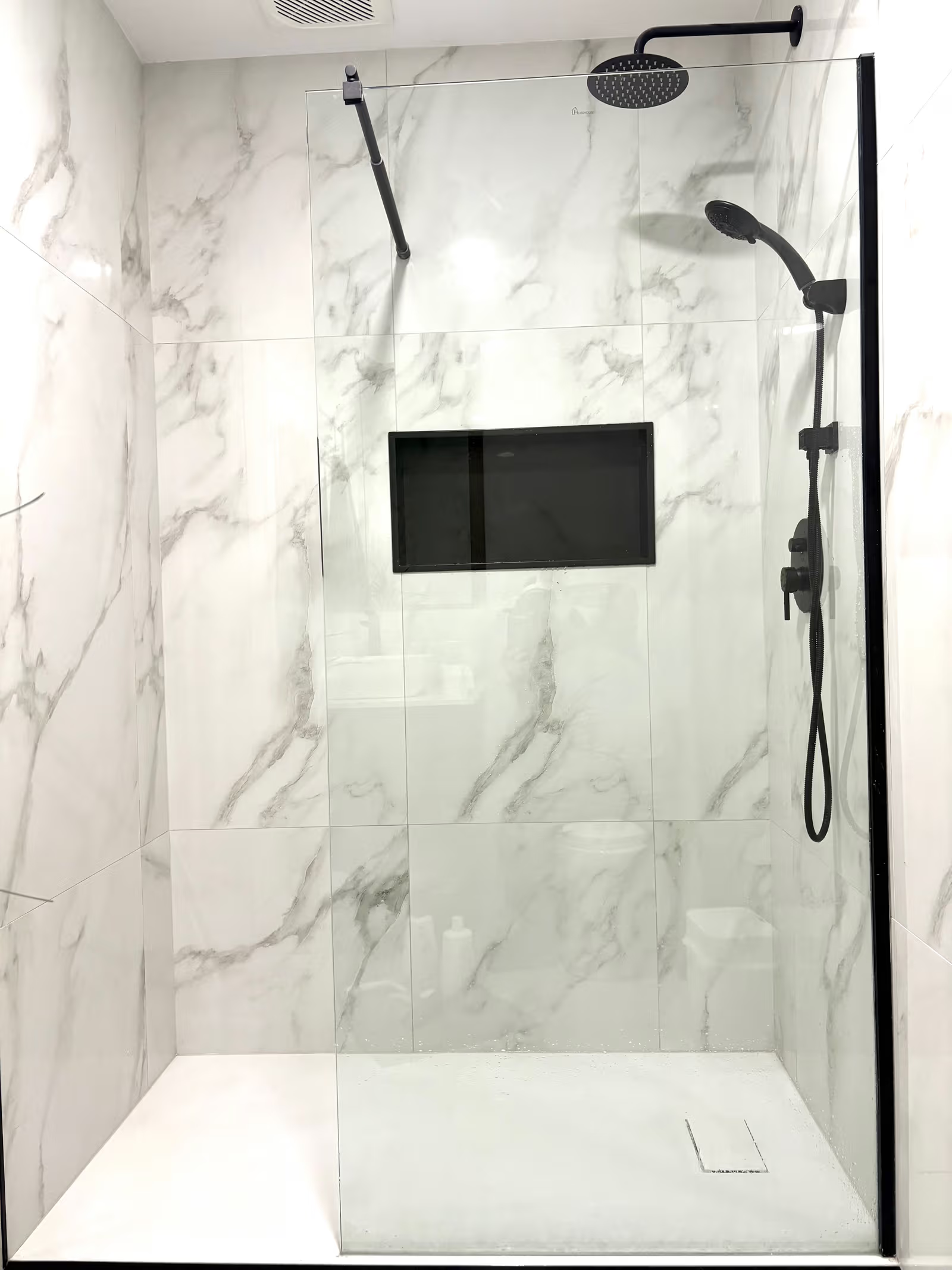 Douche italienne moderne avec grandes tuiles effet marbre, paroi de verre, niche murale noire et robinetterie noire avec pommeau pluie.