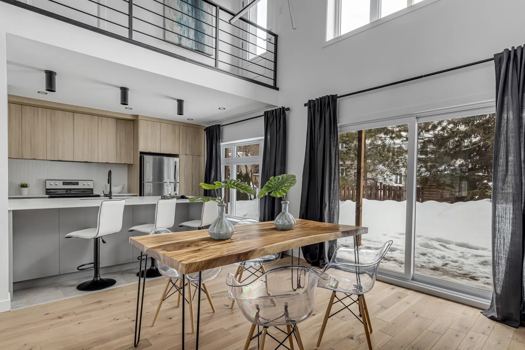 &nbsp;Modern dining room with a wooden table, transparent chairs, and an open kitchen with light wood cabinets.
