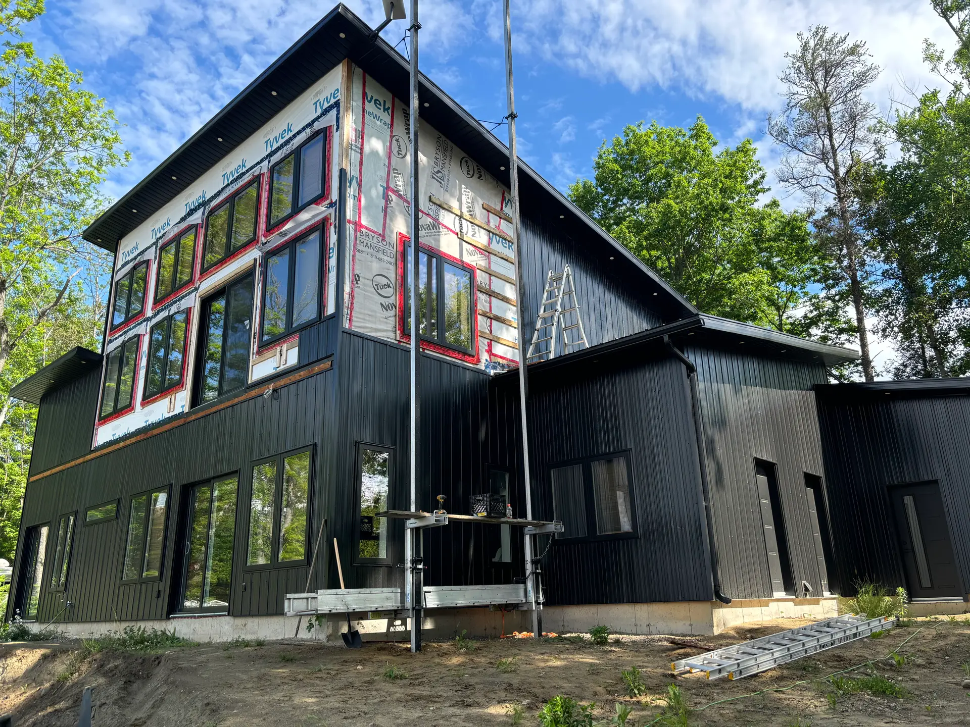 Maison en forêt avec revêtement extérieur vertical et horizontal beige, grandes fenêtres noires et toiture en pente.