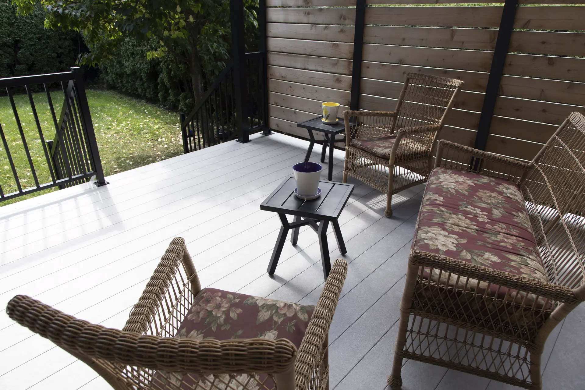 Terrasse extérieure moderne avec plancher gris, garde-corps noir, mur d’intimité en bois horizontal et mobilier de jardin en rotin avec coussins floraux.