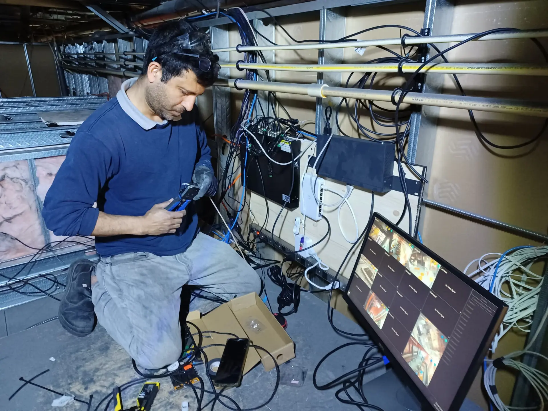 Technician configuring a security camera system with network wiring, electronic boxes, and a video monitoring screen.