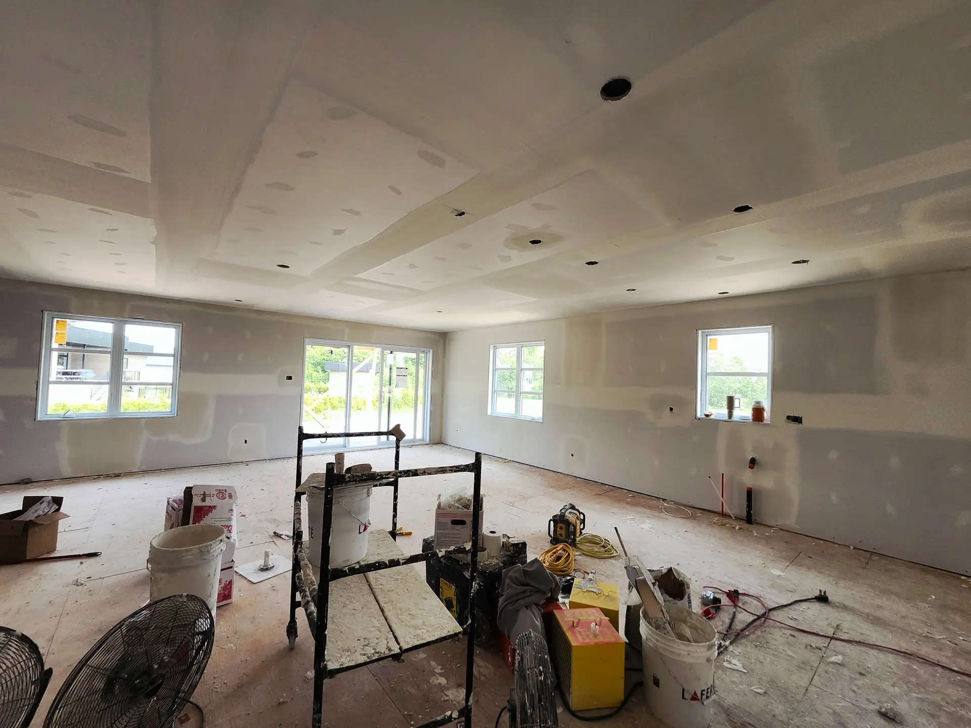Large room under renovation with taped drywall walls and ceiling, recessed light openings, scaffolding, and plastering tools.