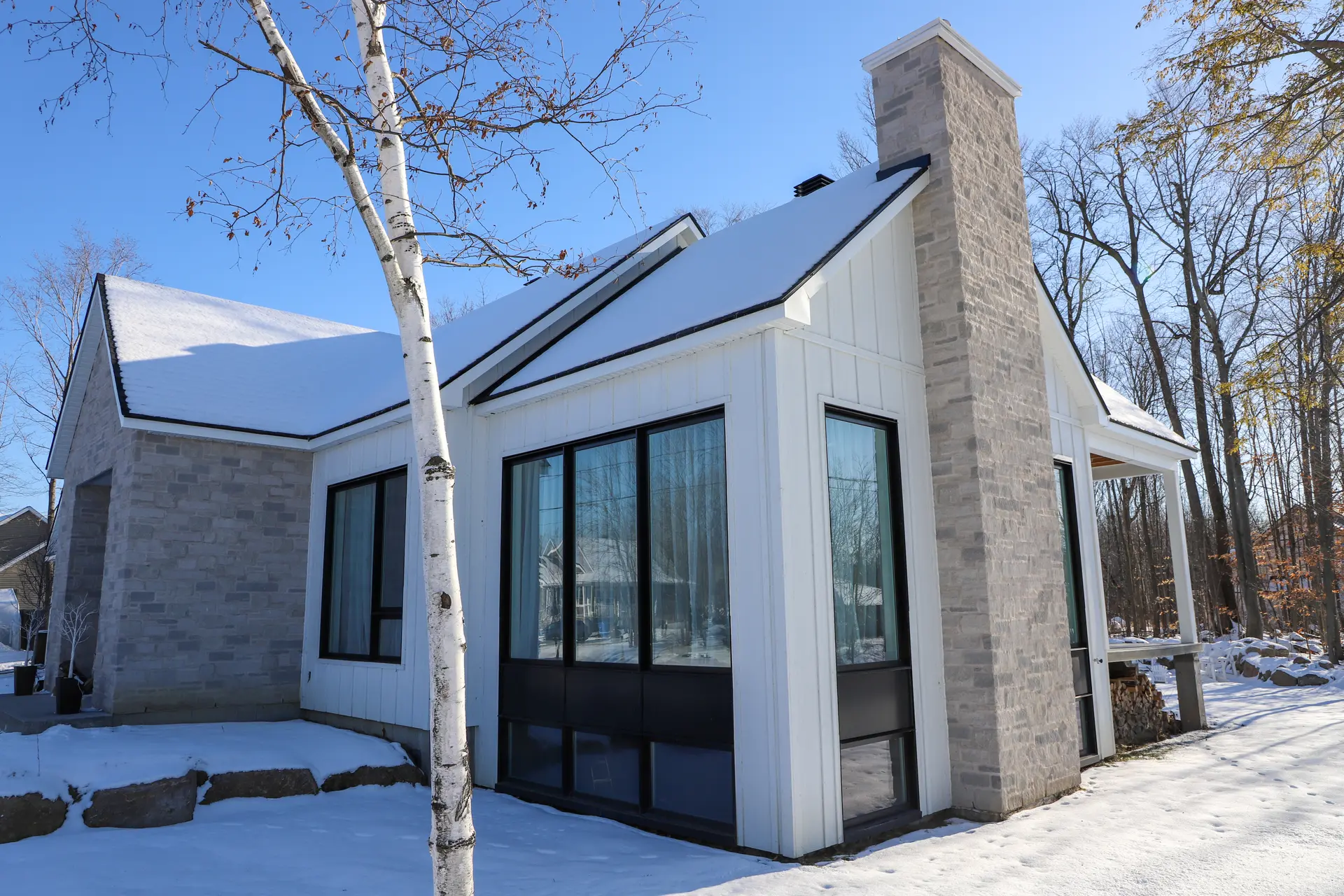 Maison moderne en hiver avec revêtement en bois blanc et pierre, grandes fenêtres et cheminée en pierre dans un environnement boisé.