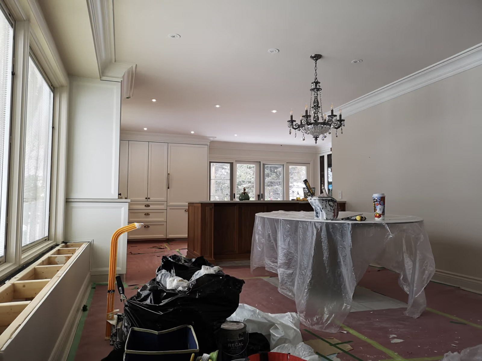 Kitchen and dining room under renovation with white cabinets, wood island, chandelier, windows, protective sheets, and painting supplies.