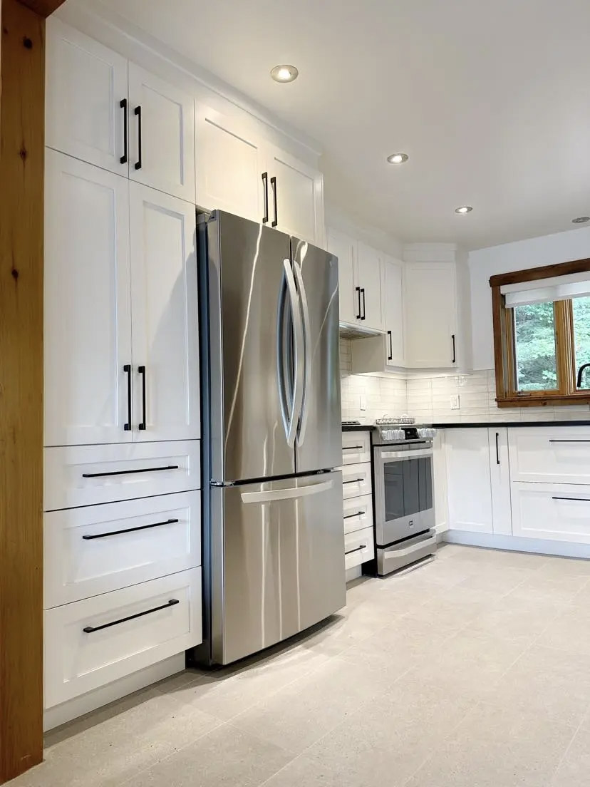 Cuisine moderne et lumineuse avec armoires blanches pleine hauteur, réfrigérateur en acier inoxydable, poignées noires, dosseret en tuiles métro blanches, plancher en céramique beige et cadrage de fenêtre en bois naturel.