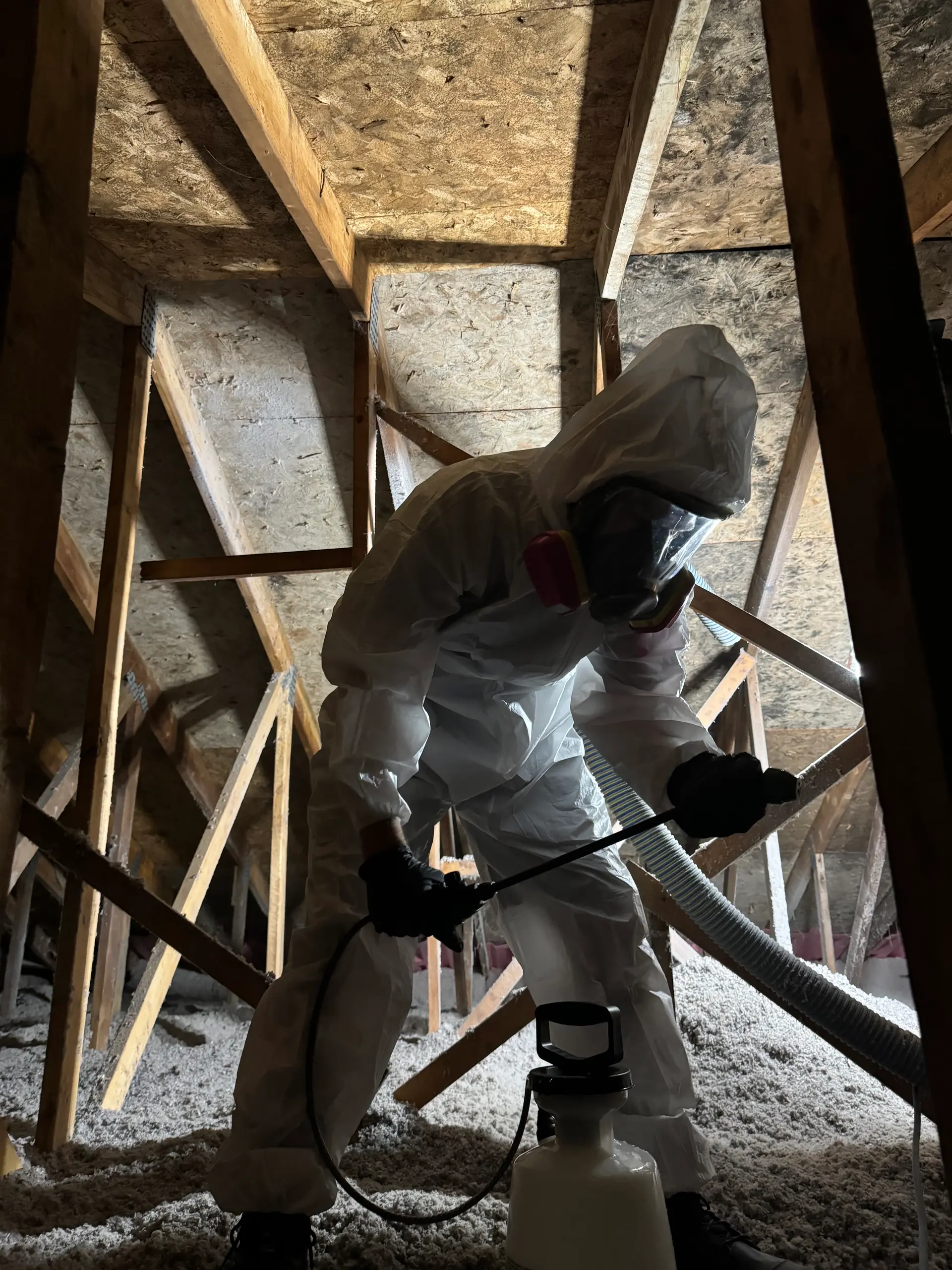 Technicien en combinaison de protection appliquant un traitement de décontamination dans un grenier avec isolation soufflée