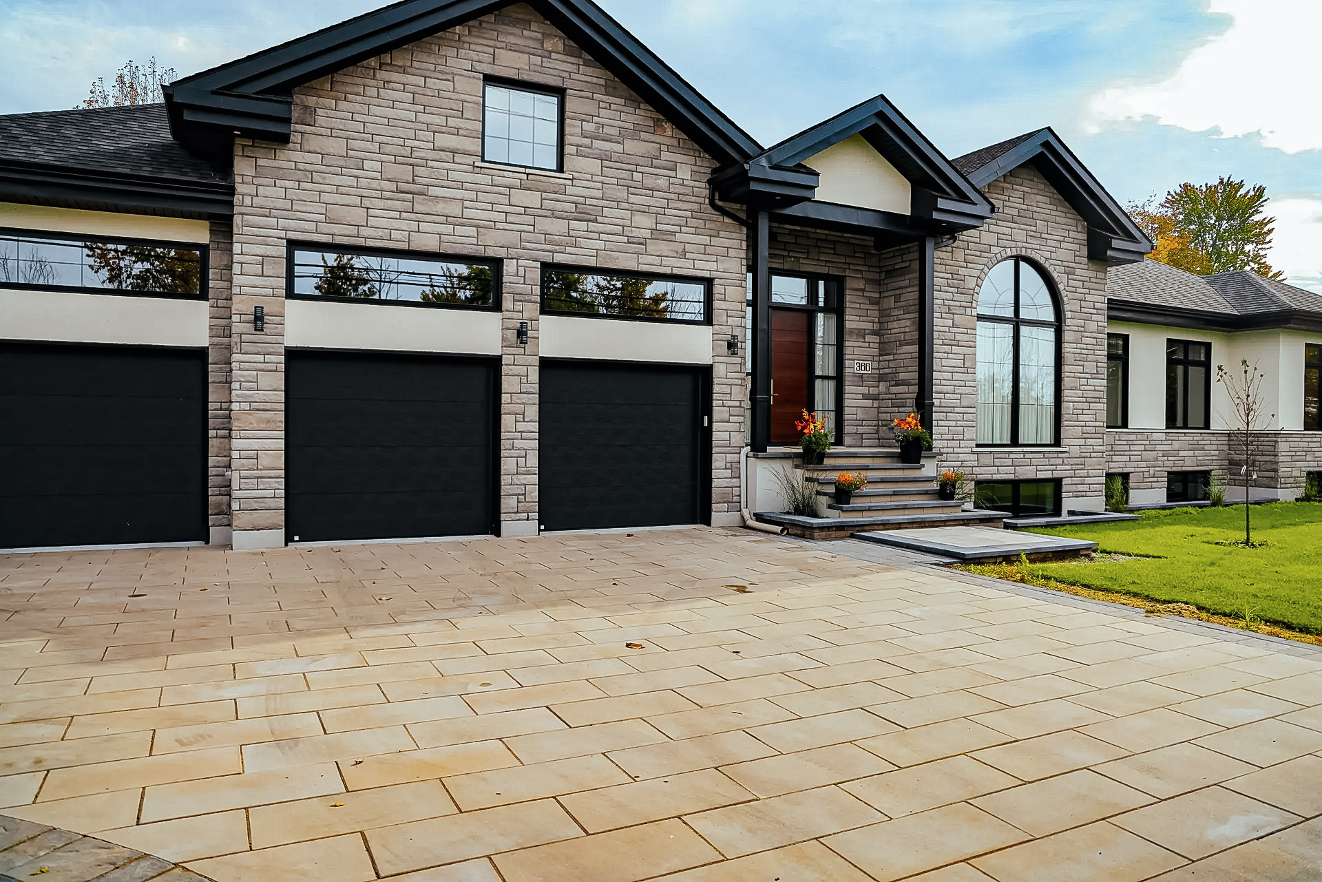 &nbsp;Maison moderne avec façade en pierre, trois portes de garage noires et entrée aménagée avec un grand pavé uni beige, entourée d'un jardin verdoyant.