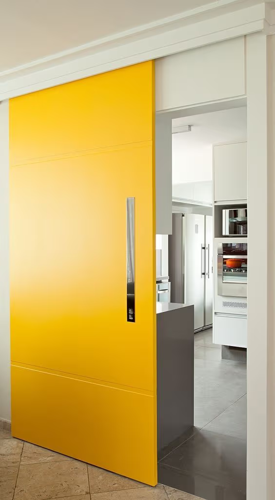Porte coulissante jaune moderne avec poignée verticale en métal, séparant une cuisine contemporaine aux tons blancs et gris.